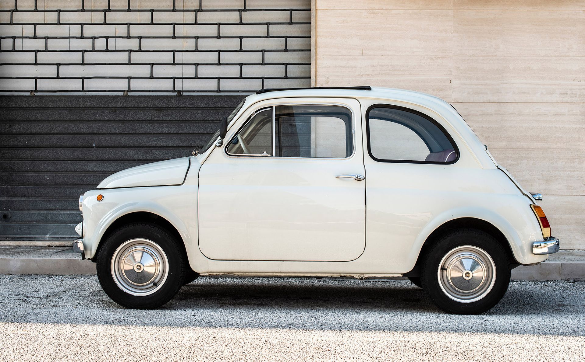 White vintage Fiat 500 car parked on a cobblestone street, set against brick and beige walls.