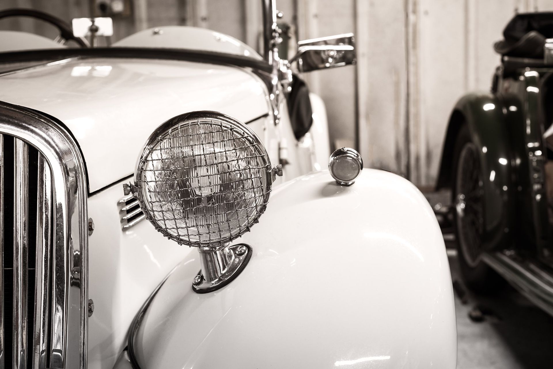 Classic white car's front with a round headlight and another car's fender to the right. Monochrome.