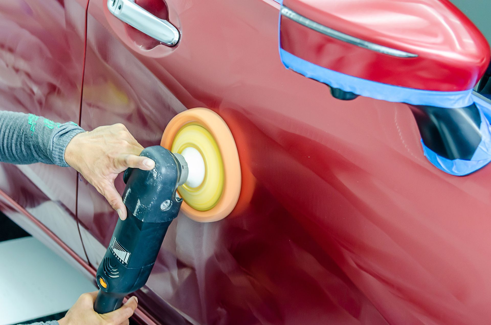 Person using a power polisher on a red car door. Masking tape protects the mirror.