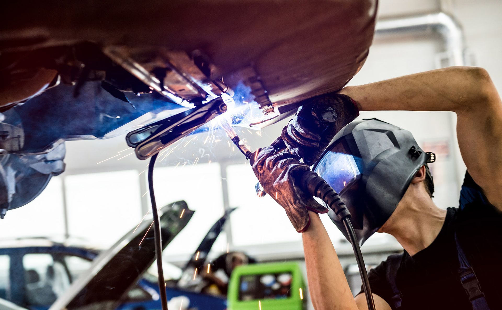 Mechanic welding a car part with sparks flying in a brightly lit workshop.