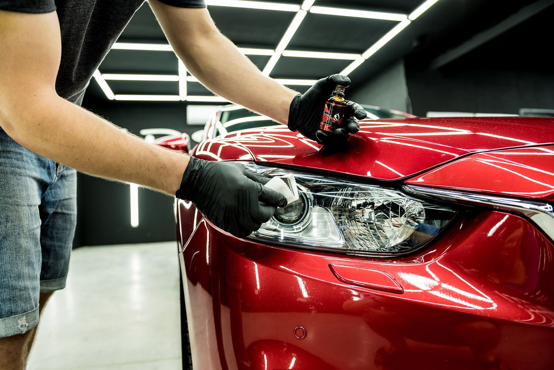 Person wearing gloves applying car wax to a red car's headlight.