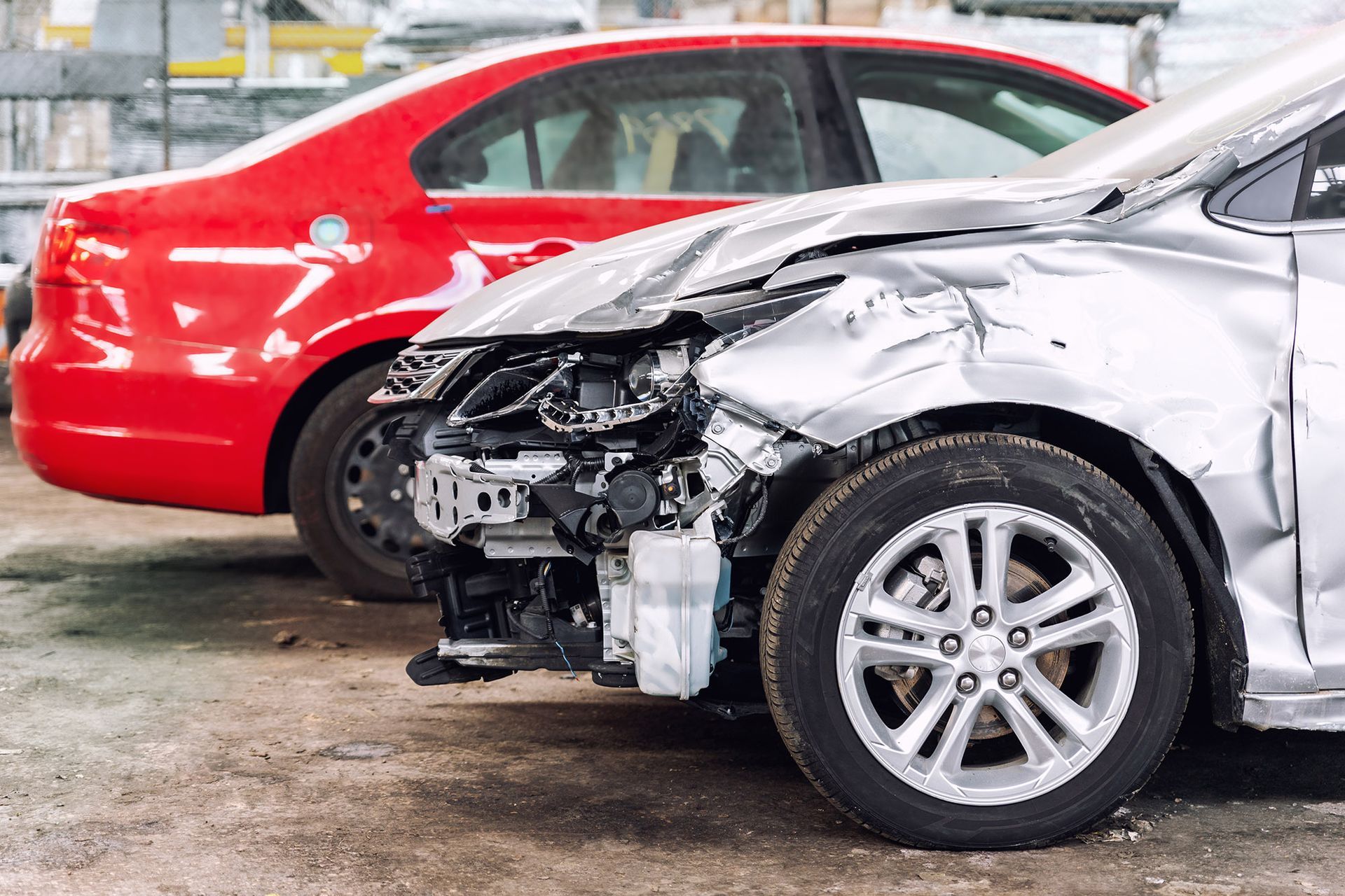 Damaged silver car with front-end collision; red car in background.