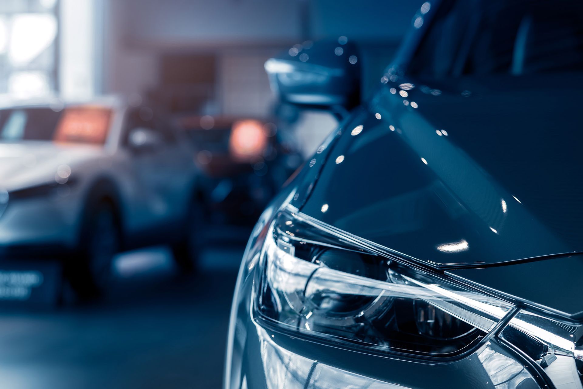 Close-up of a blue car's headlight in a showroom, with another car blurred in the background.