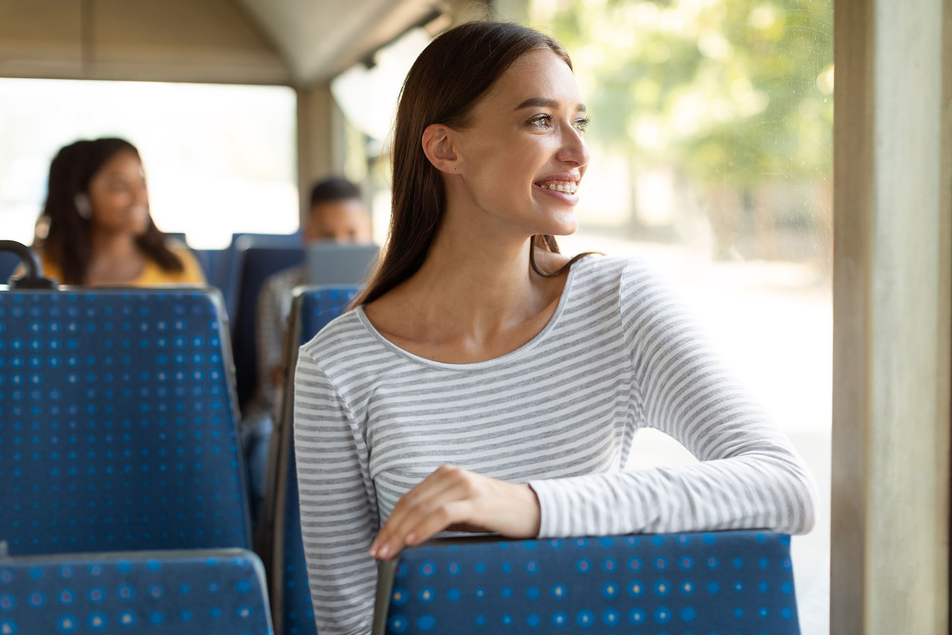 Woman on a bus, smiling and looking out window, resting hand on seat.