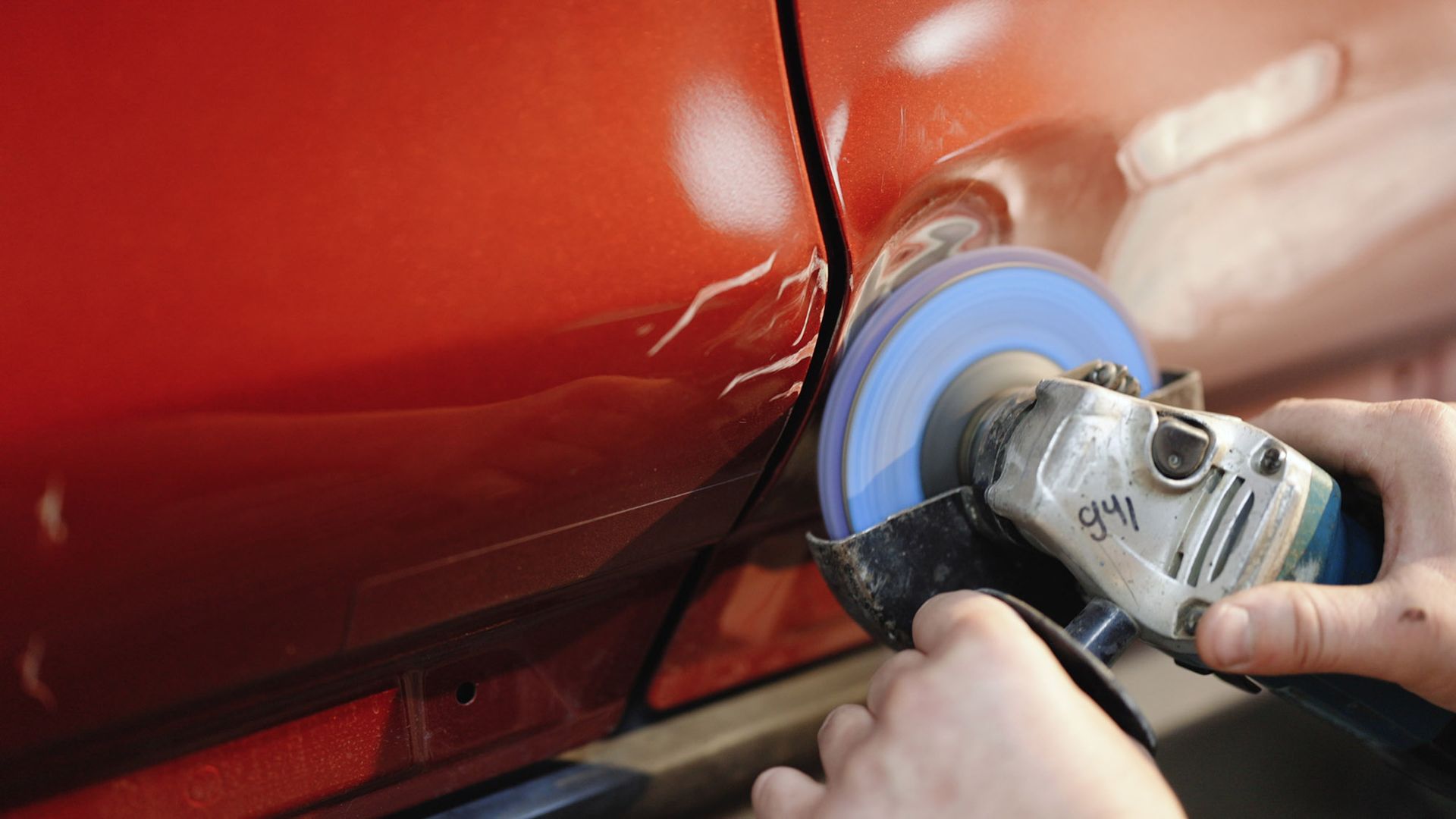 Person uses a grinder to sand the scratched surface of a red car door.