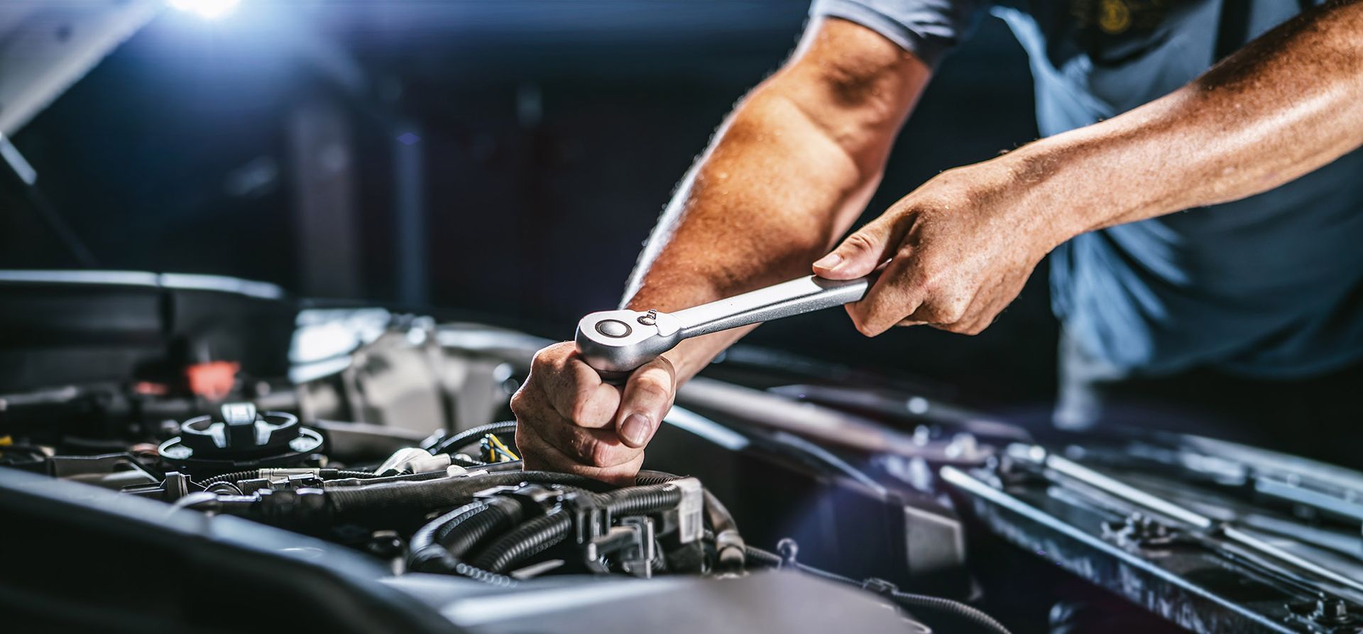 Mechanic working on a car engine with a wrench. Dark garage setting, bright light overhead.