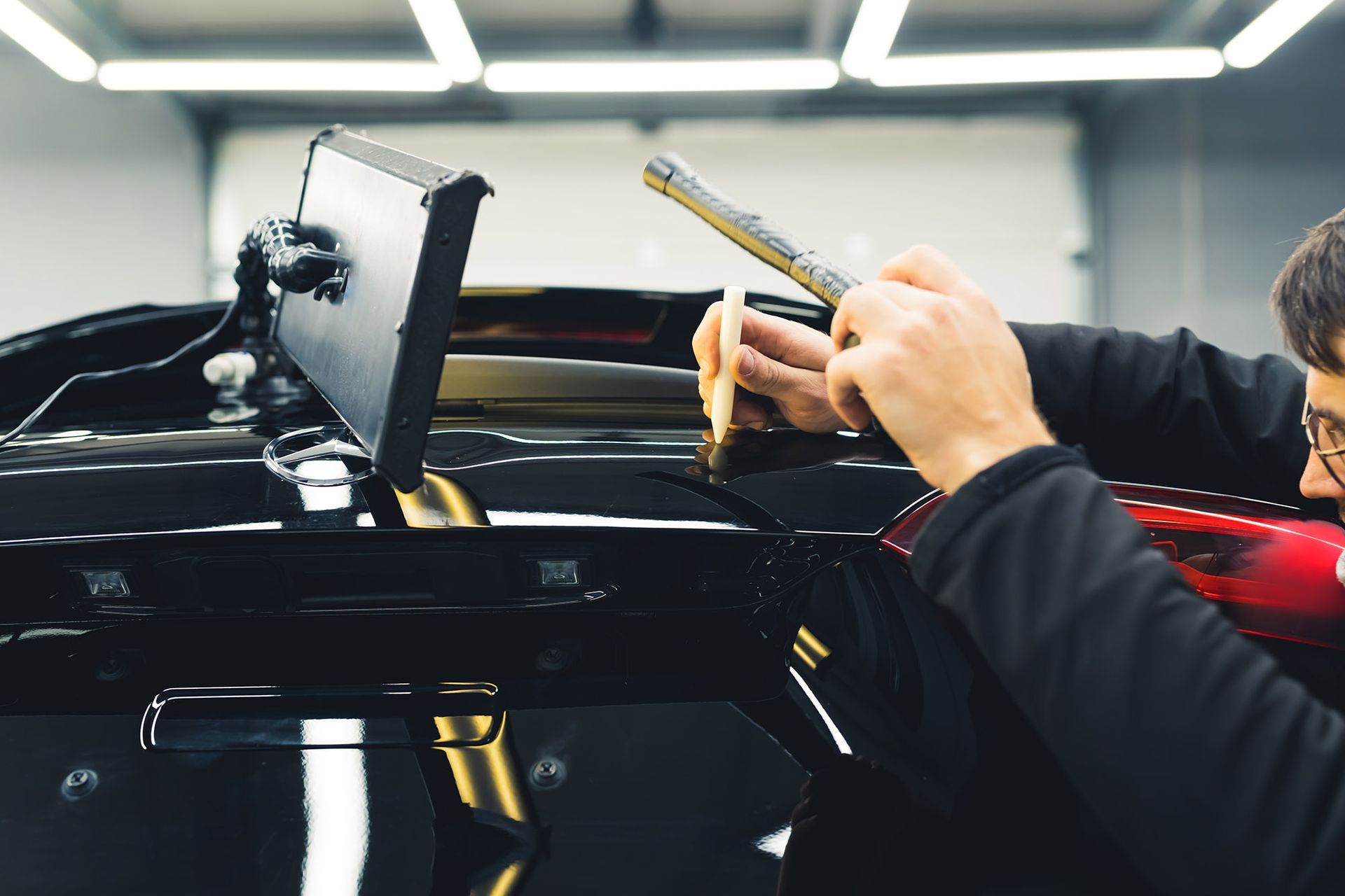 Person using tools to apply a film on a black car in a well-lit workshop.
