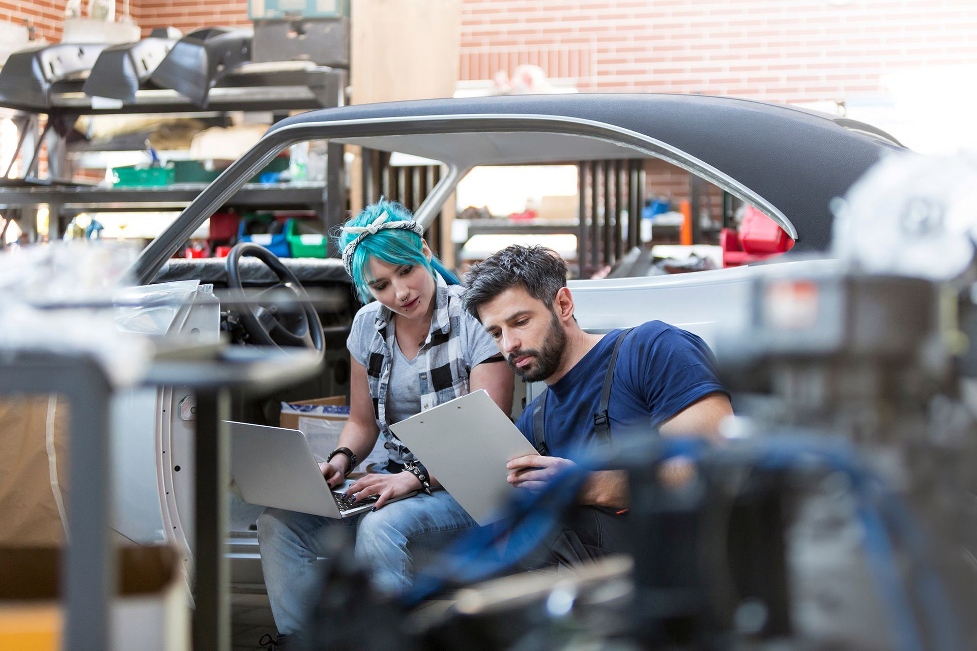 Two people, one with teal hair and a clipboard, work on a classic car, looking at a laptop.