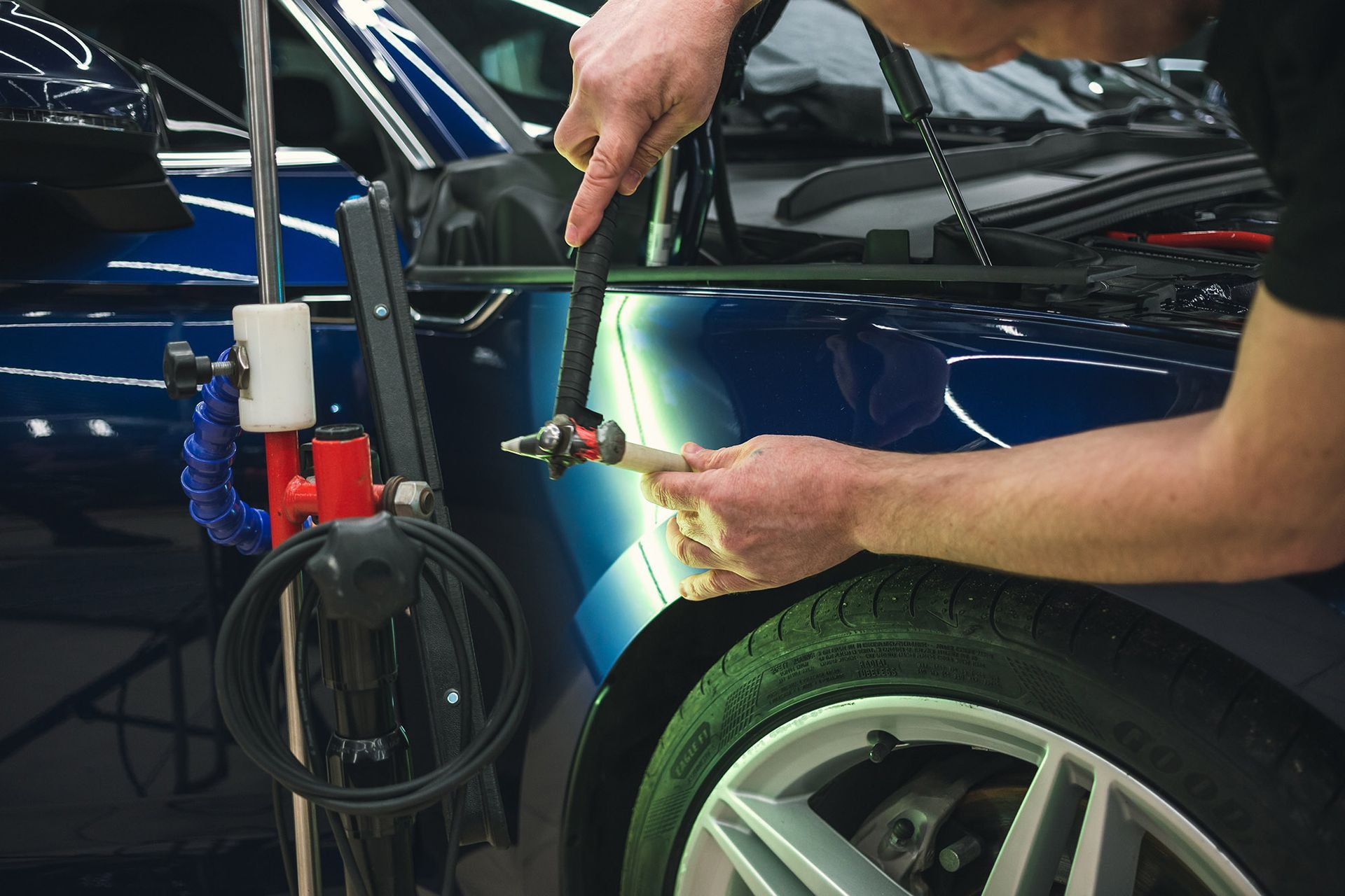 Person using tools to remove a dent from a blue car's fender near a tire.