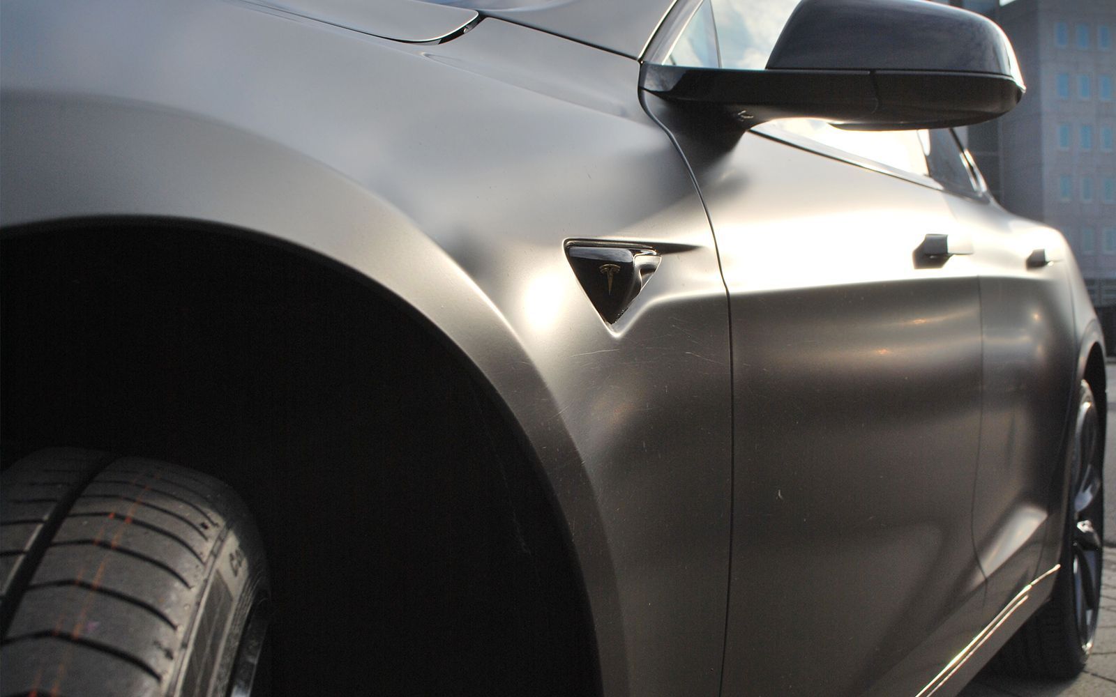 Matte gray Tesla car, front quarter view, tire visible, side mirror, bright light reflection.