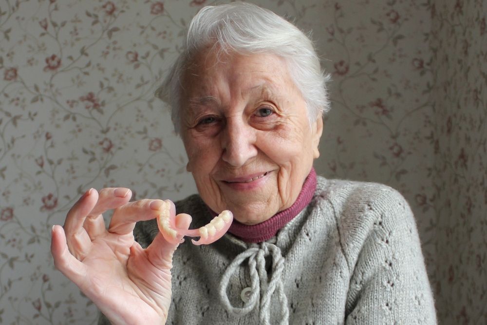 Elderly woman smiling, holding a set of dentures in front of her, indoors.