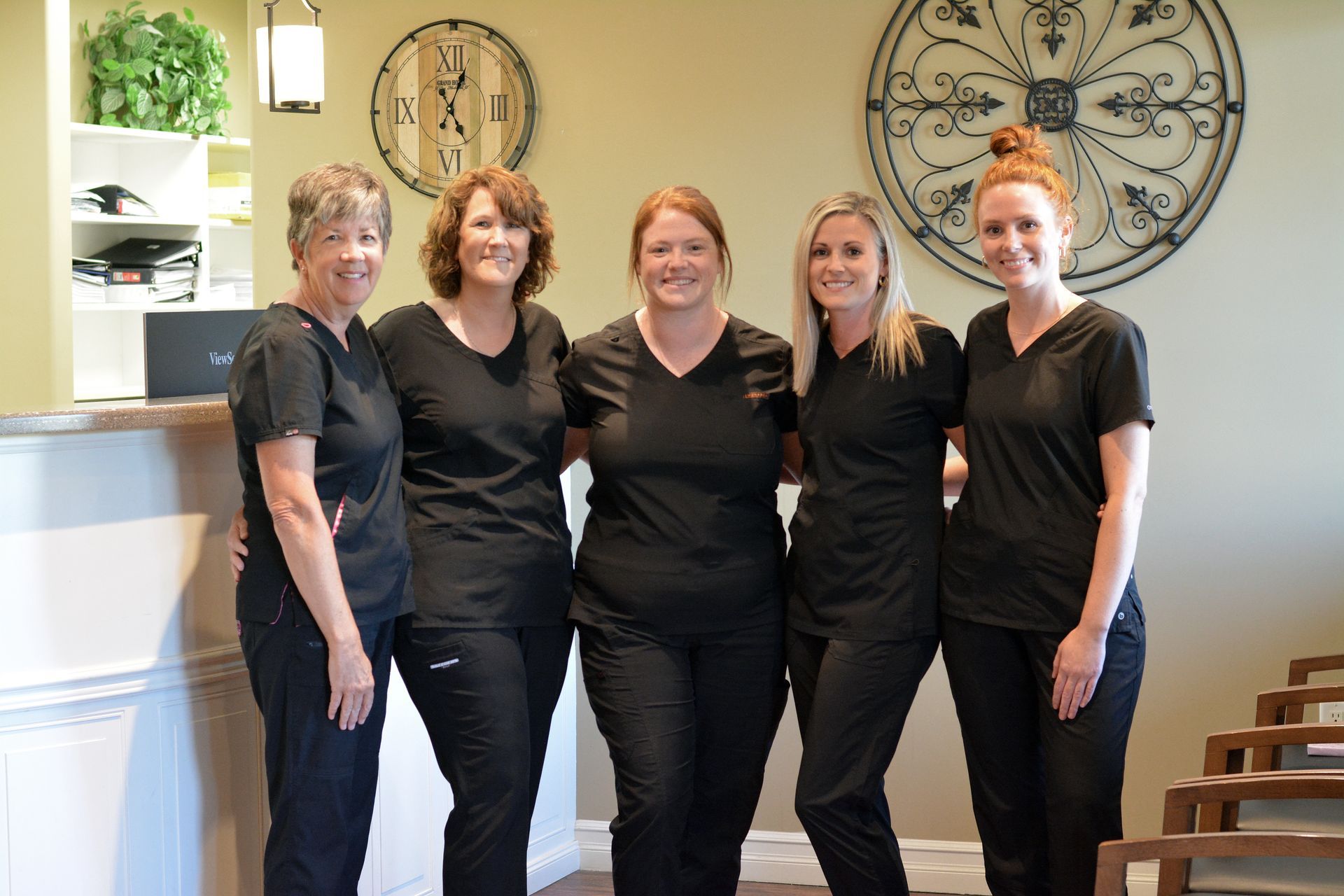 Five women in black scrubs standing in a dental office, smiling.