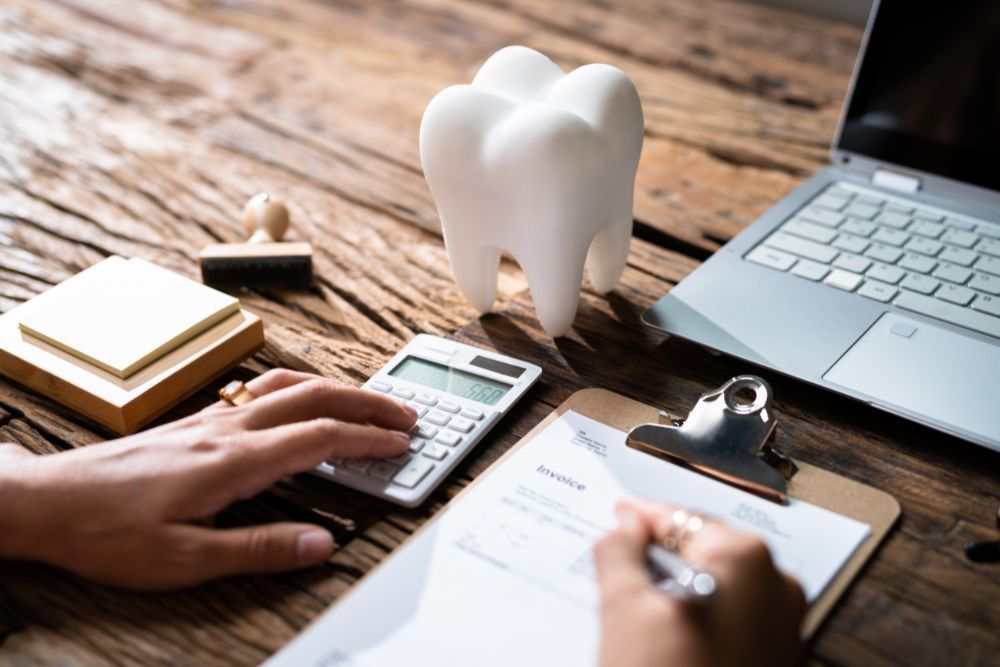 Hands calculating dental bill with calculator and pen, tooth model, clipboard, and laptop on a wooden desk.