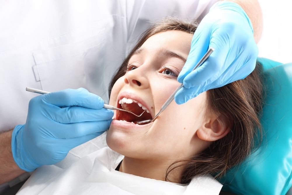Girl with mouth open at dentist, being examined. Dentist wearing gloves holds instruments.