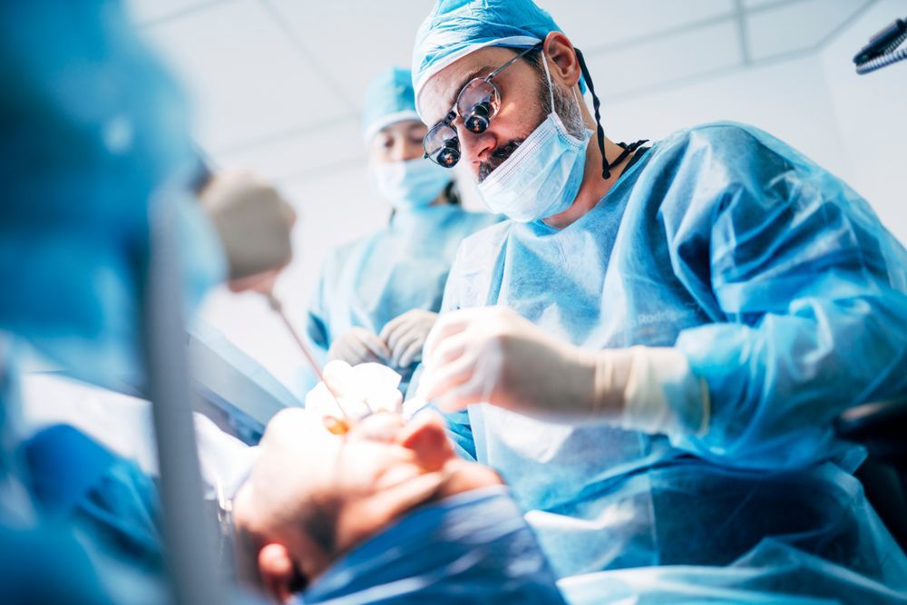 Surgeons in blue scrubs operating on a patient's face in a brightly lit operating room.