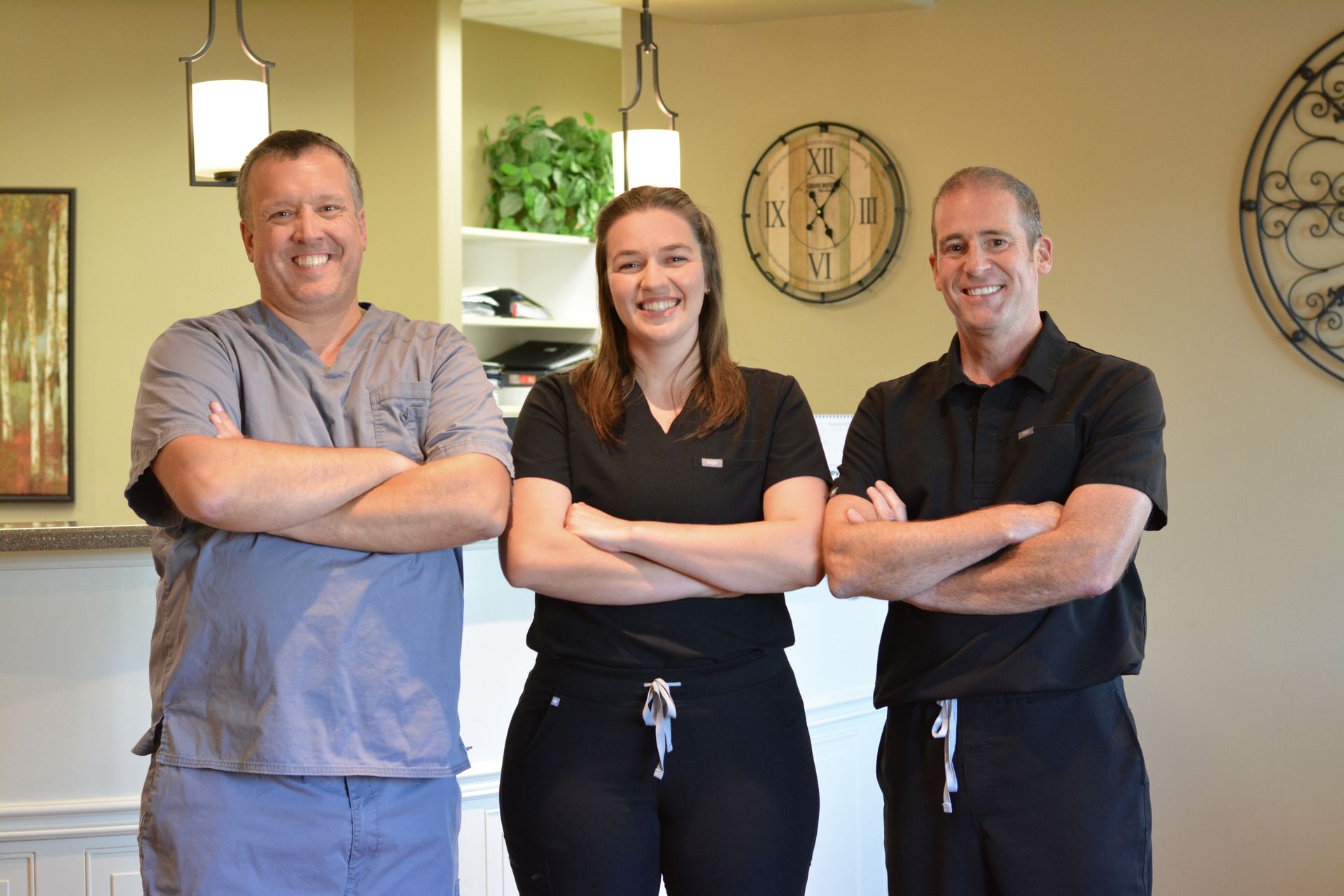 Three medical professionals, arms crossed, smiling. Two men and one woman in scrubs. Neutral background.