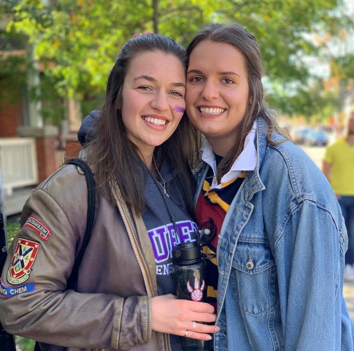 Two smiling women in a sunny outdoor setting. One wears a jacket, the other a denim jacket and tie.