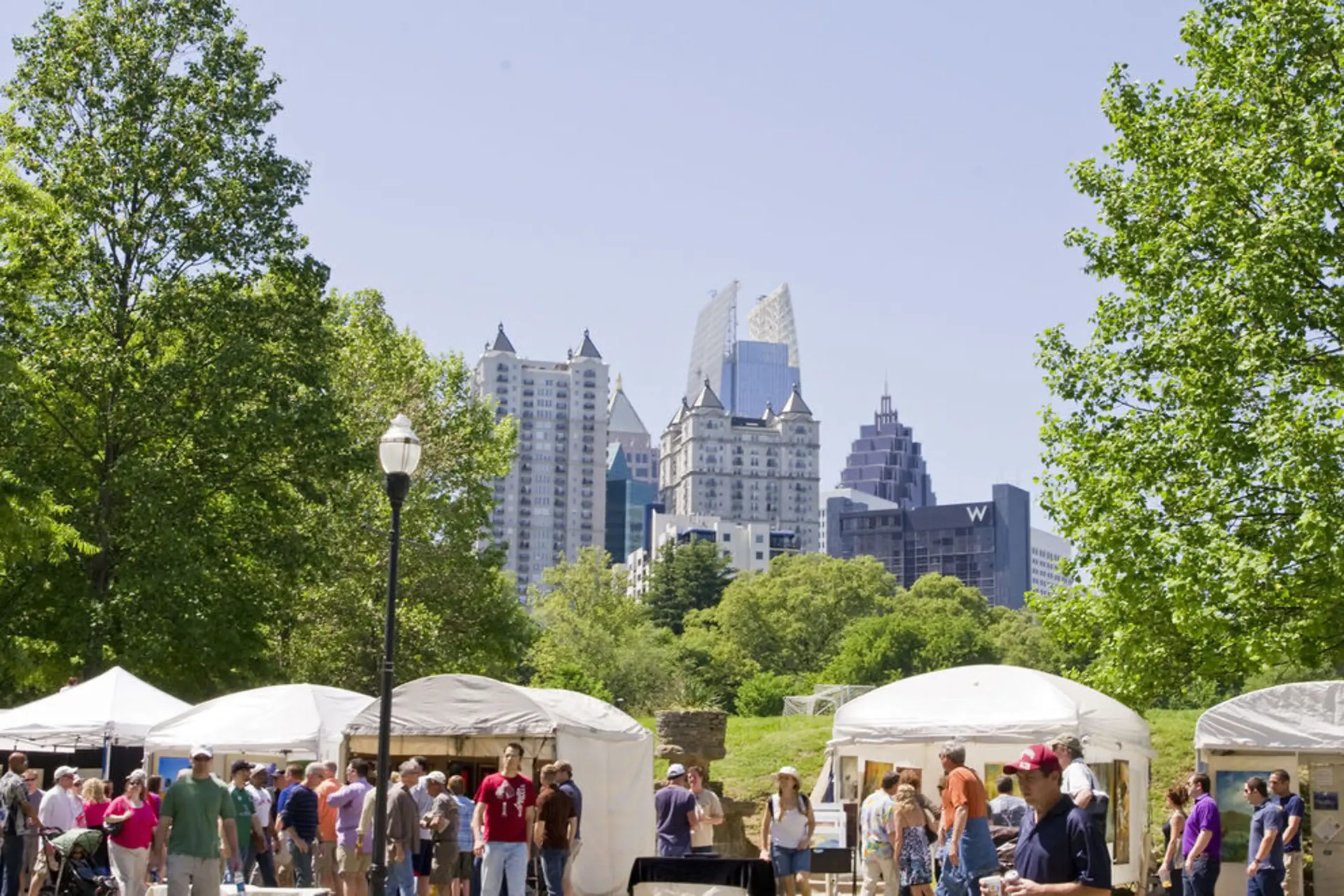Crowded park market with white tents and a city skyline behind green trees