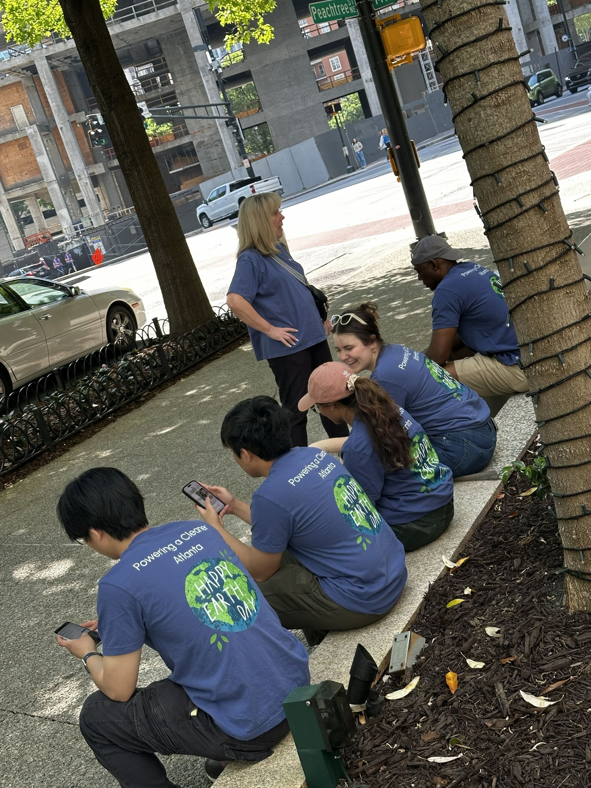 People sitting on a tree-ringed sidewalk planter, chatting in blue shirts near a street.