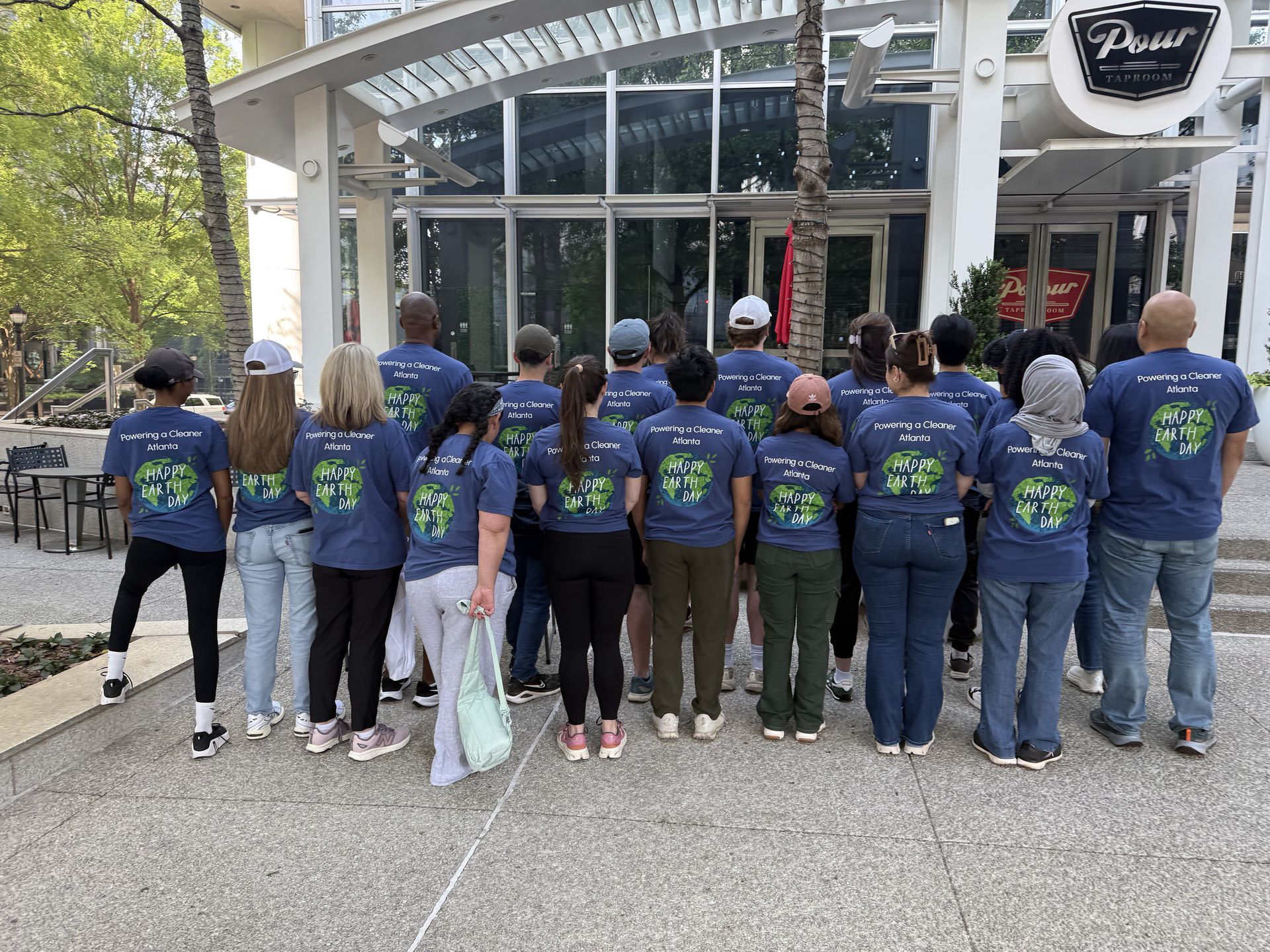 Group in matching blue shirts standing outside a glass-fronted building, seen from behind.