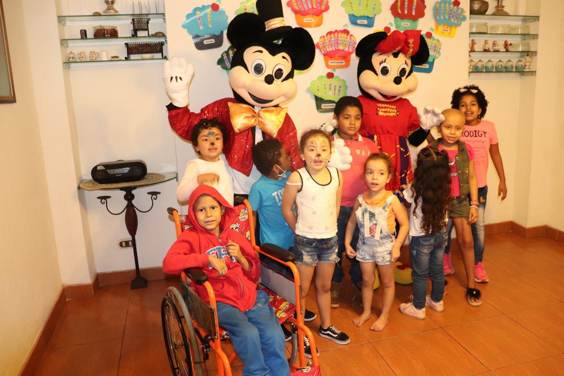 Niños posan con Mickey y Minnie Mouse en una habitación decorada con pastelitos. Un niño usa silla de ruedas.
