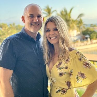 A man and a woman are posing for a picture together . the woman is wearing a yellow floral dress.