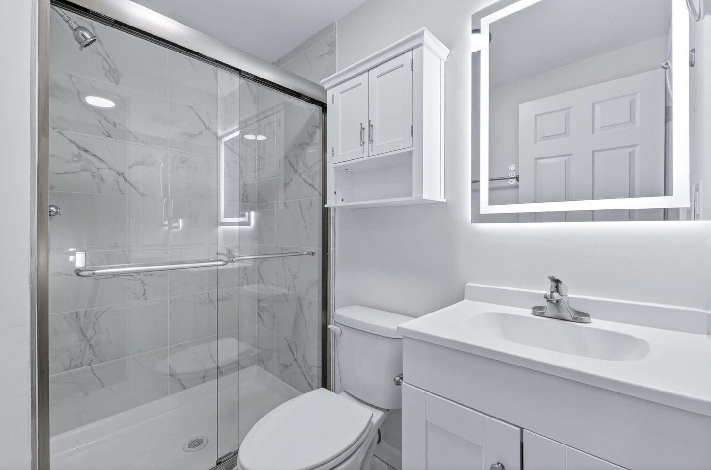 Bright white bathroom with glass shower, toilet, sink, and illuminated mirror