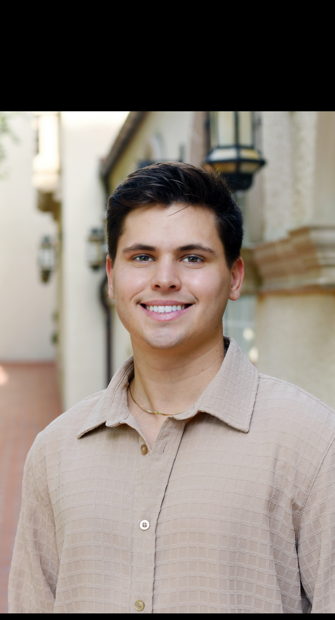Young man with a warm smile, wearing a tan button-up shirt, outdoors in a light-colored, ornate building.