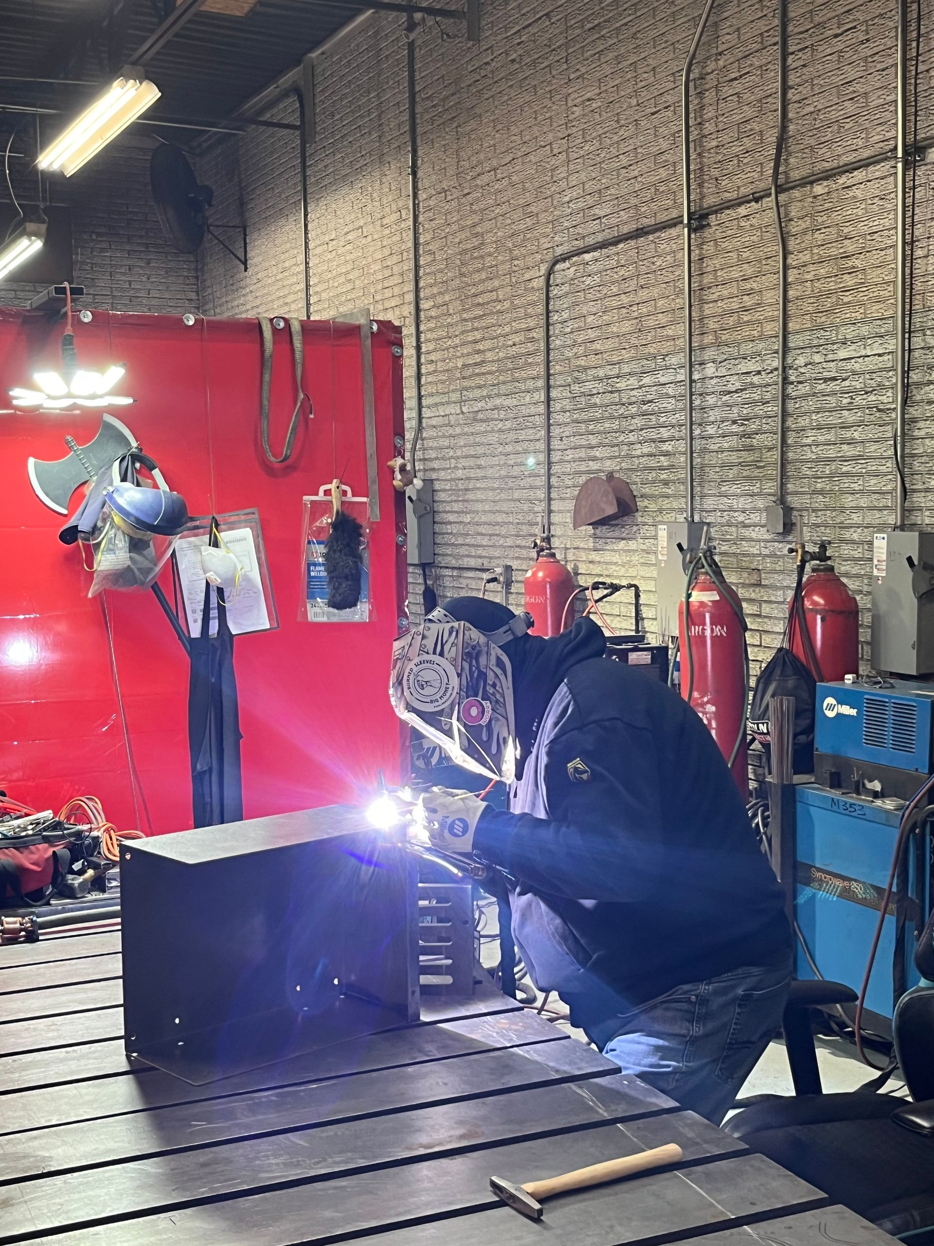 Welder in a workshop, wearing a helmet, welding a metal box. Brick wall backdrop, red tank, blue welding machine.