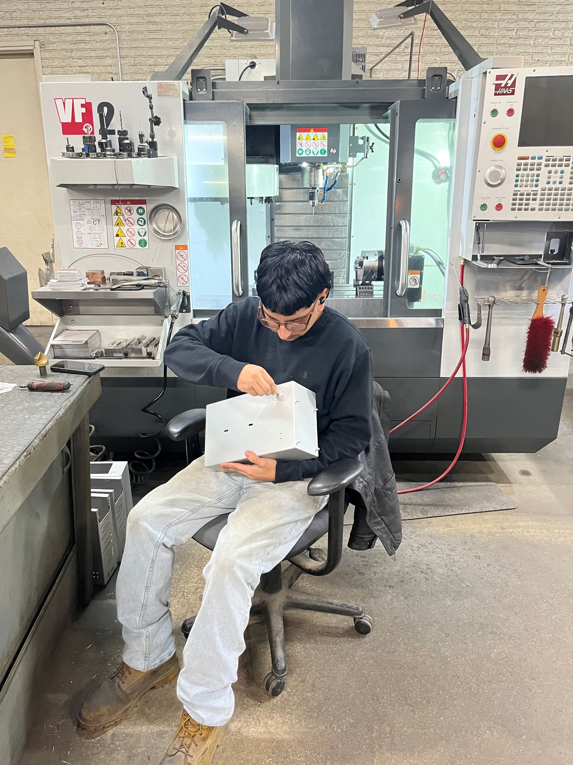 Man inspecting a white part, sitting in shop with CNC machine in the background.