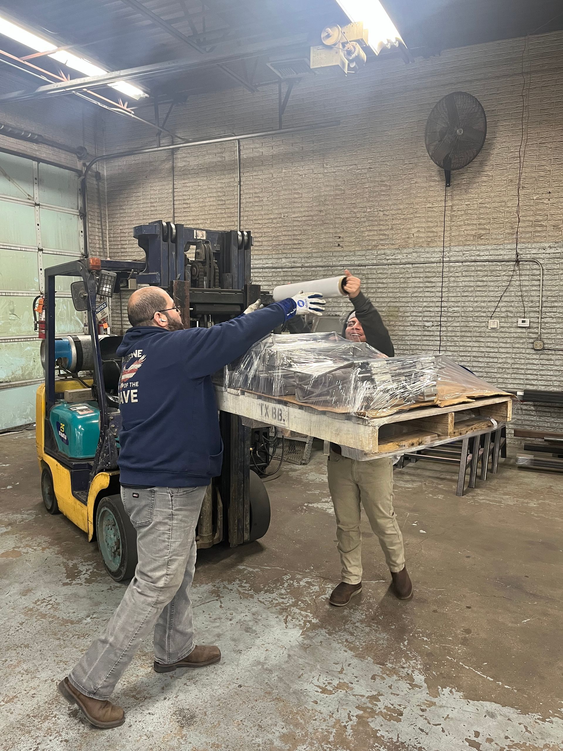 Two men loading a pallet of materials with a forklift in a warehouse; one guides, the other operates.