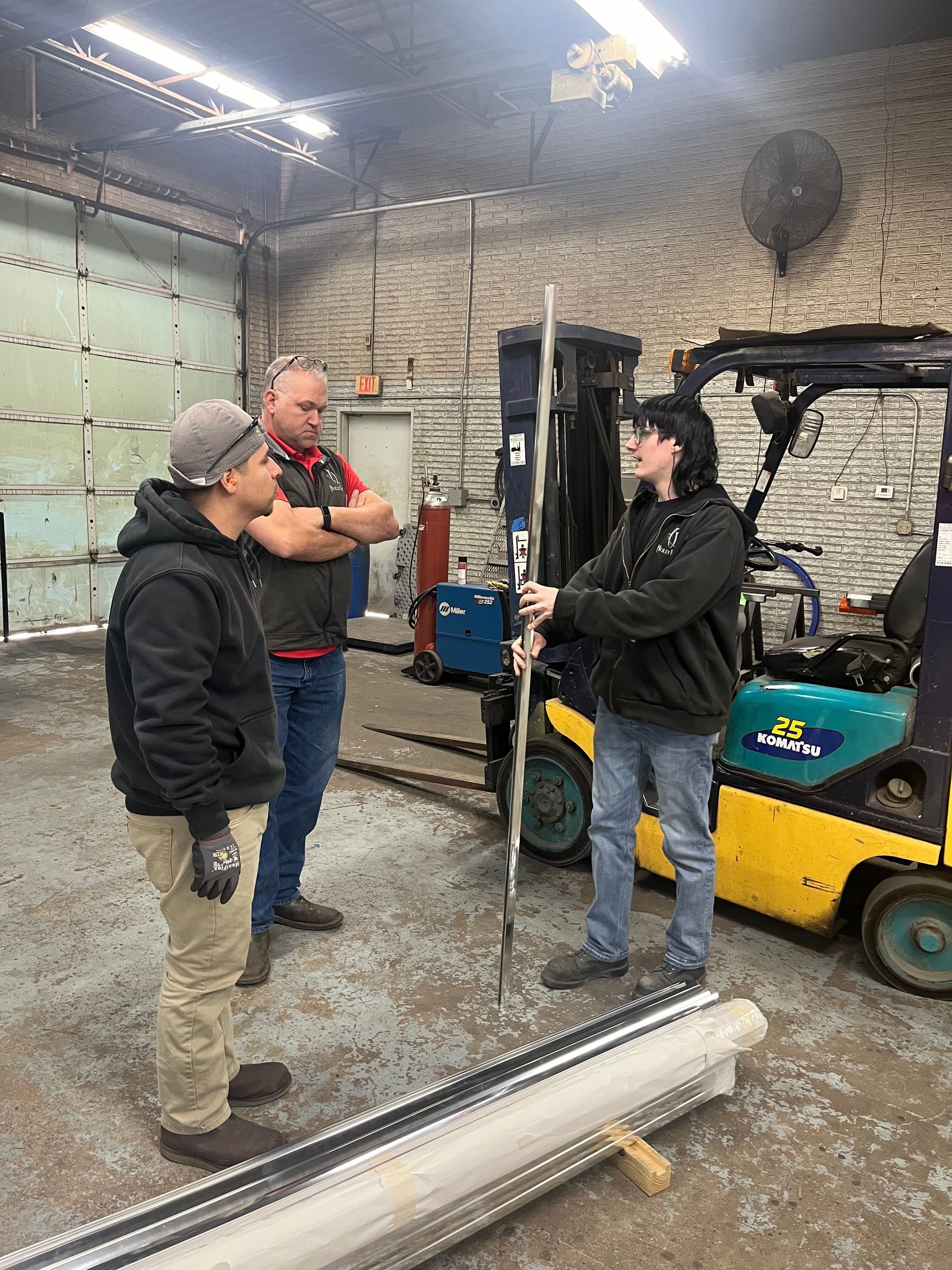 Three men discussing something, one holding a metal bar near a forklift in a workshop.
