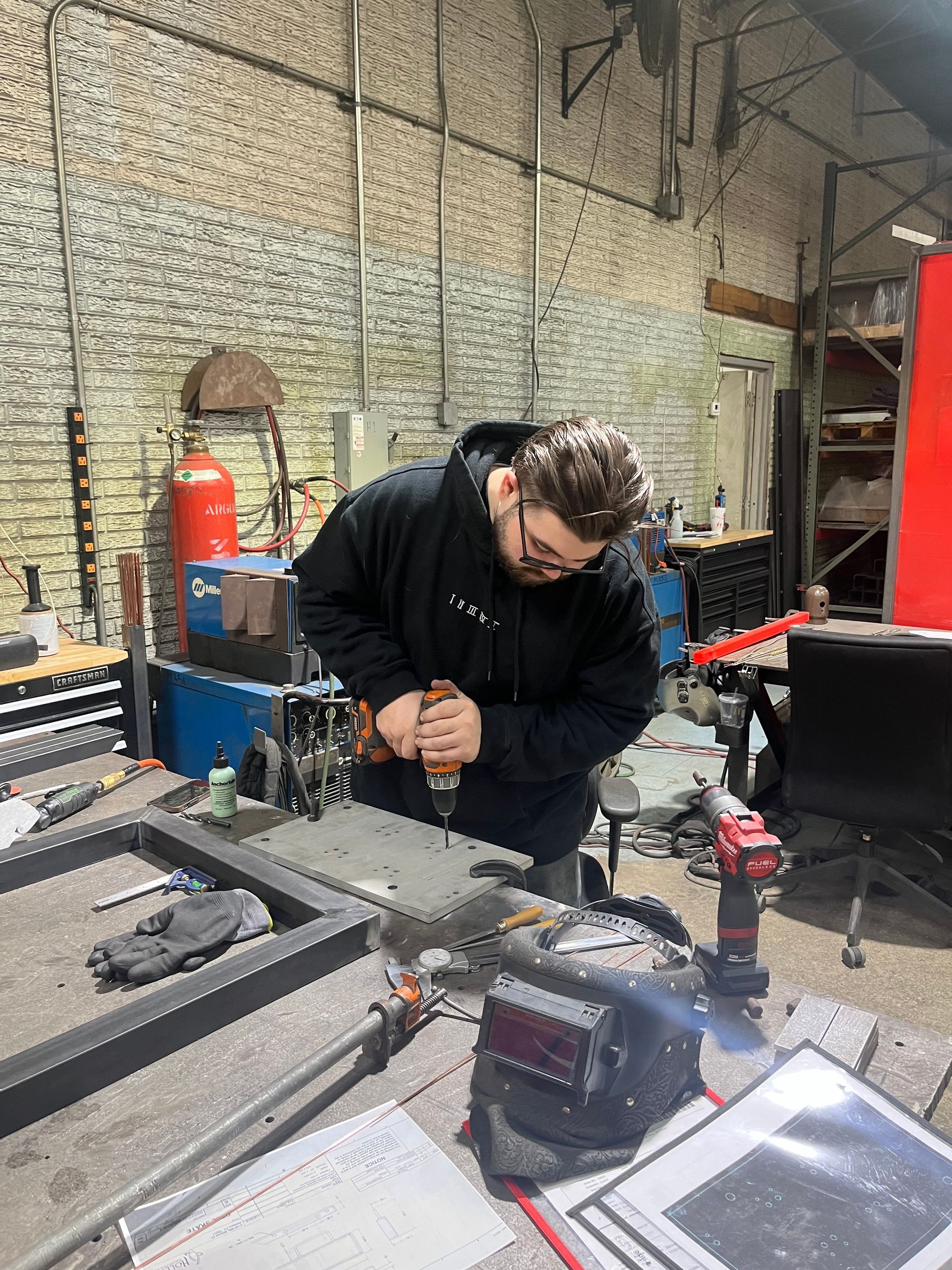 Man in workshop drills into metal frame, wearing glasses and hoodie, with tools scattered around.