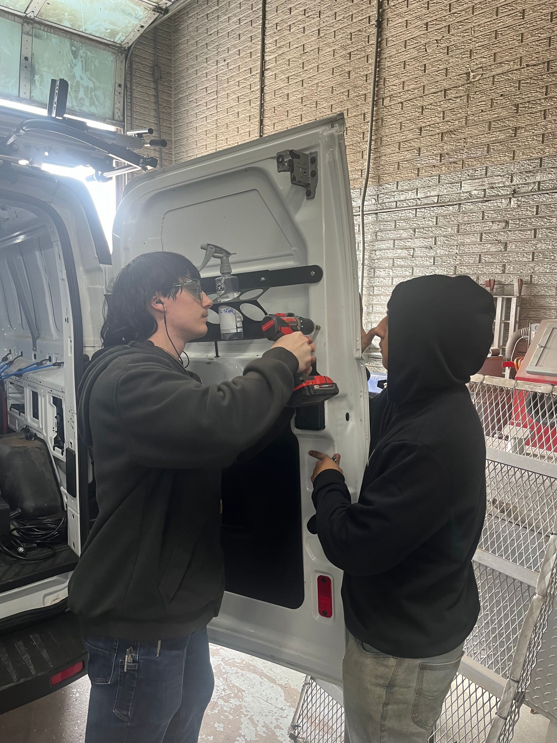 Two people work on a van door using tools inside a workshop.