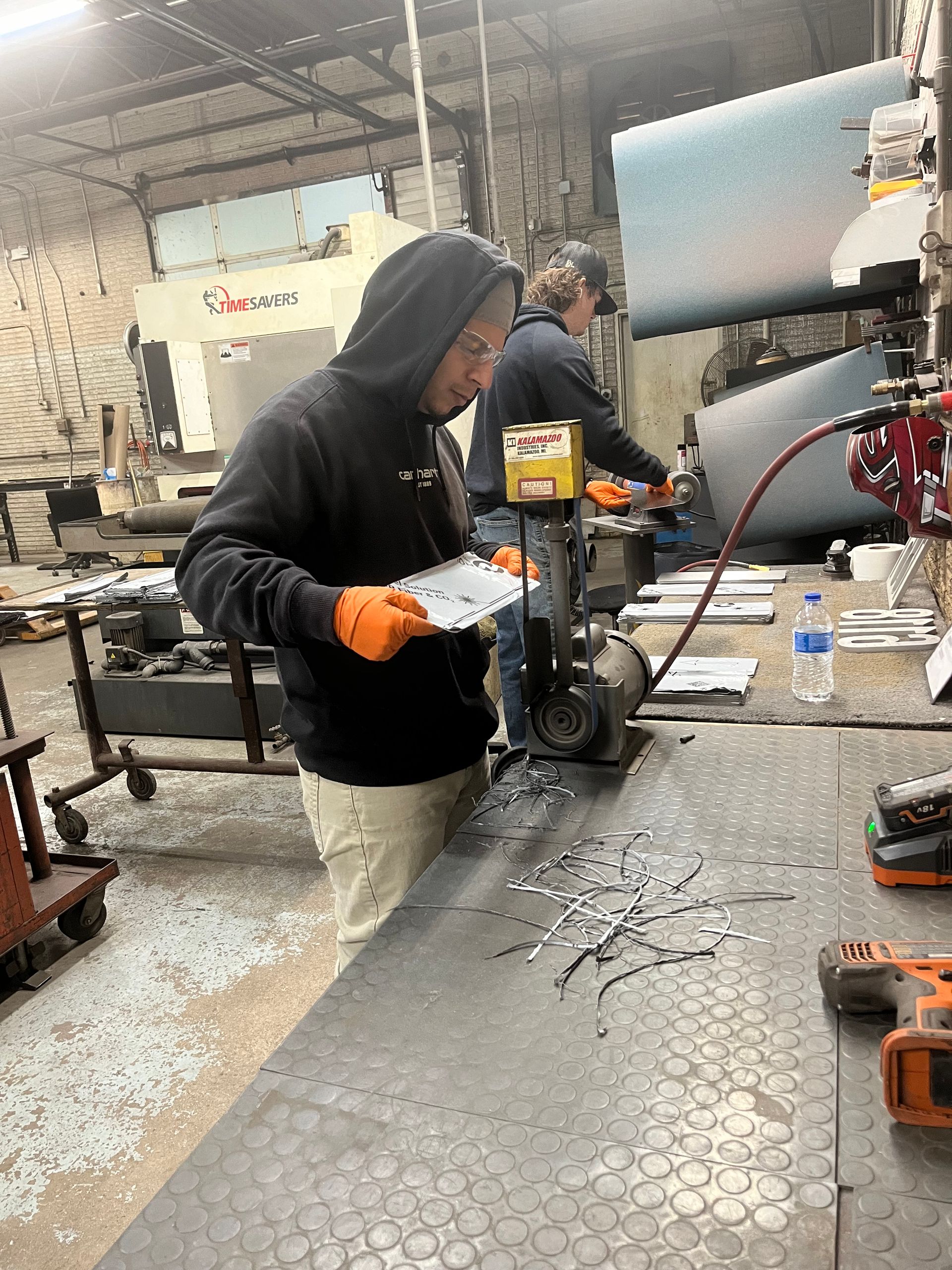 Two workers in a metal shop. One drills metal, the other uses a machine. Both wear safety gear.