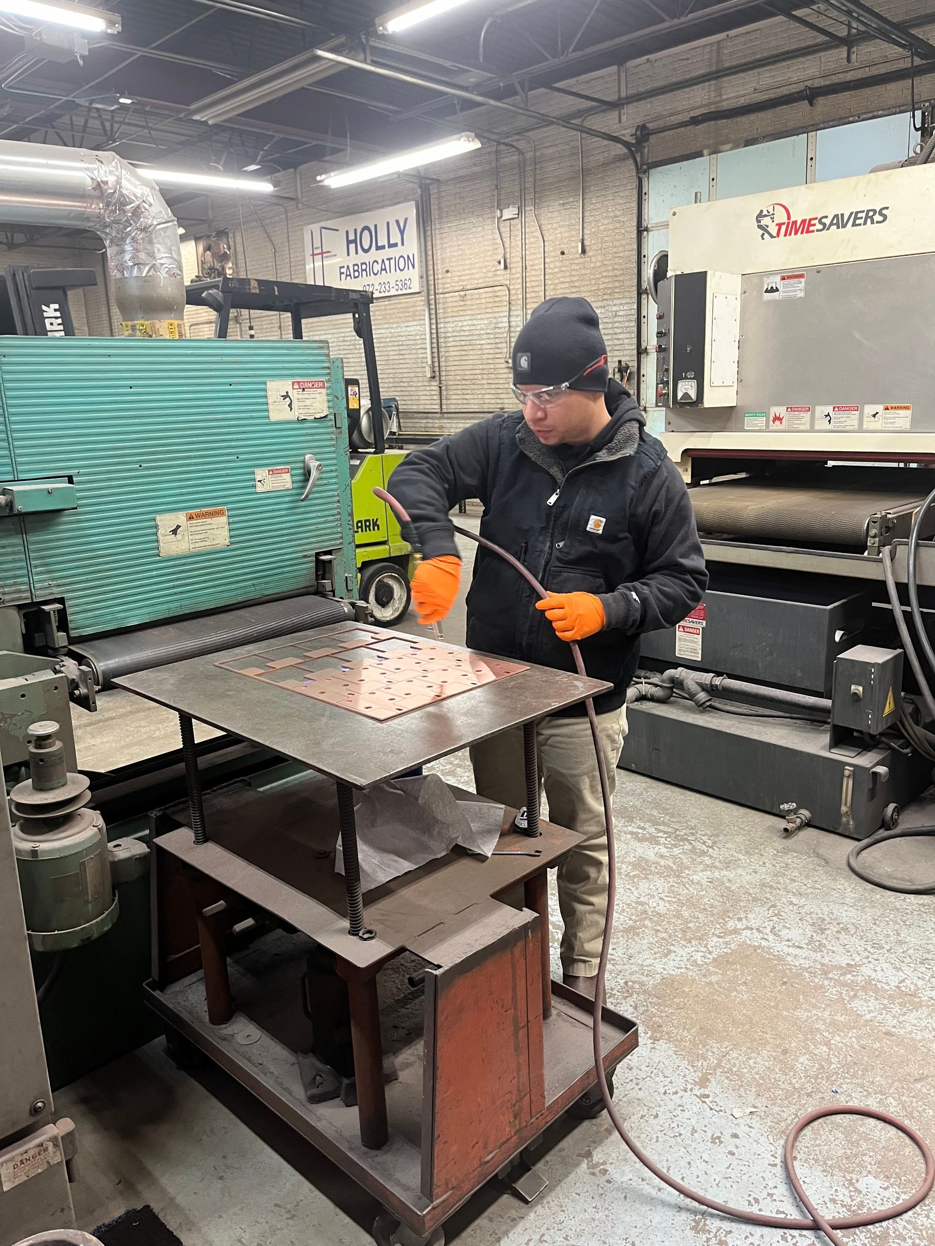 A man in a workshop uses a machine to work on a metal sheet. He wears safety glasses and gloves.