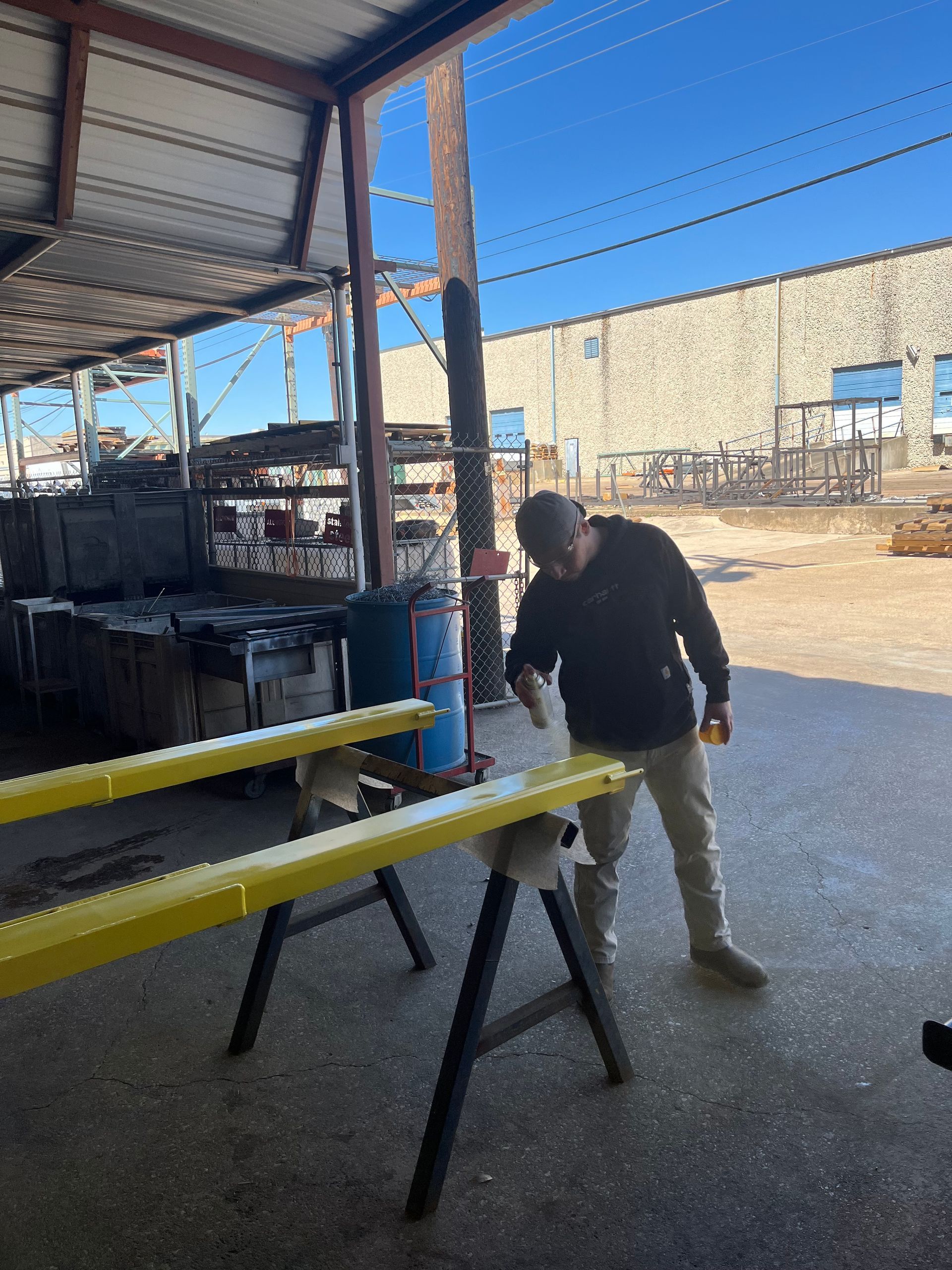 A man sprays yellow paint on metal beams supported by sawhorses, outside a warehouse.