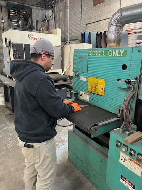 Man sanding steel with a belt sander in a workshop. He wears safety glasses, gloves, and a cap.