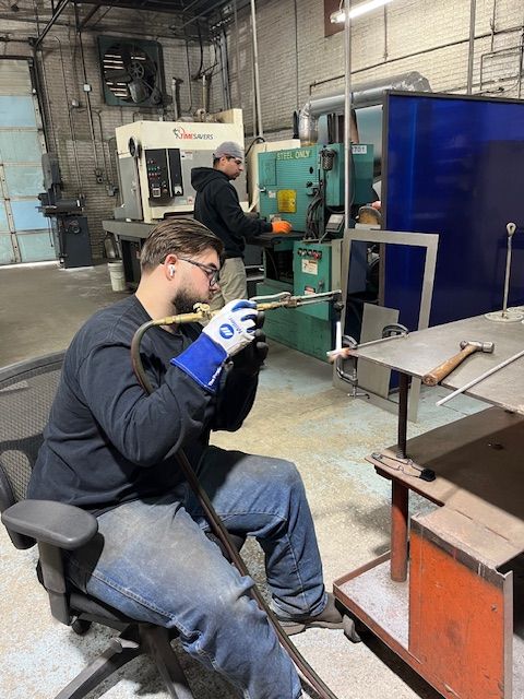 Man welding metal in a workshop, wearing safety glasses and gloves. Another man operates machinery in the background.