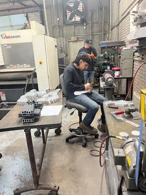 Two workers in a metal shop; one seated writing, another using a grinding wheel. Gray machinery and parts on tables.