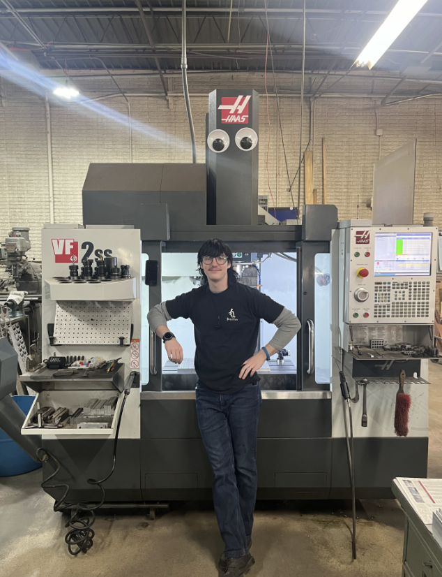 Man in a black shirt stands next to a large Haas CNC mill. He’s smiling, leaning on the machine, in a workshop.