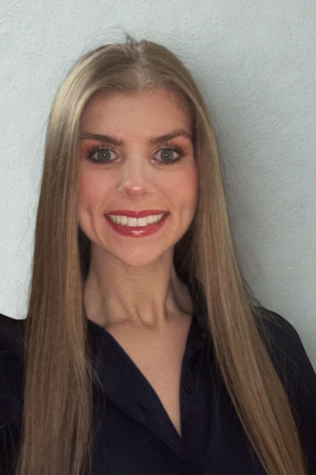 A young woman with long, curly blonde hair smiles at the camera. She wears a black top and a gray blazer.