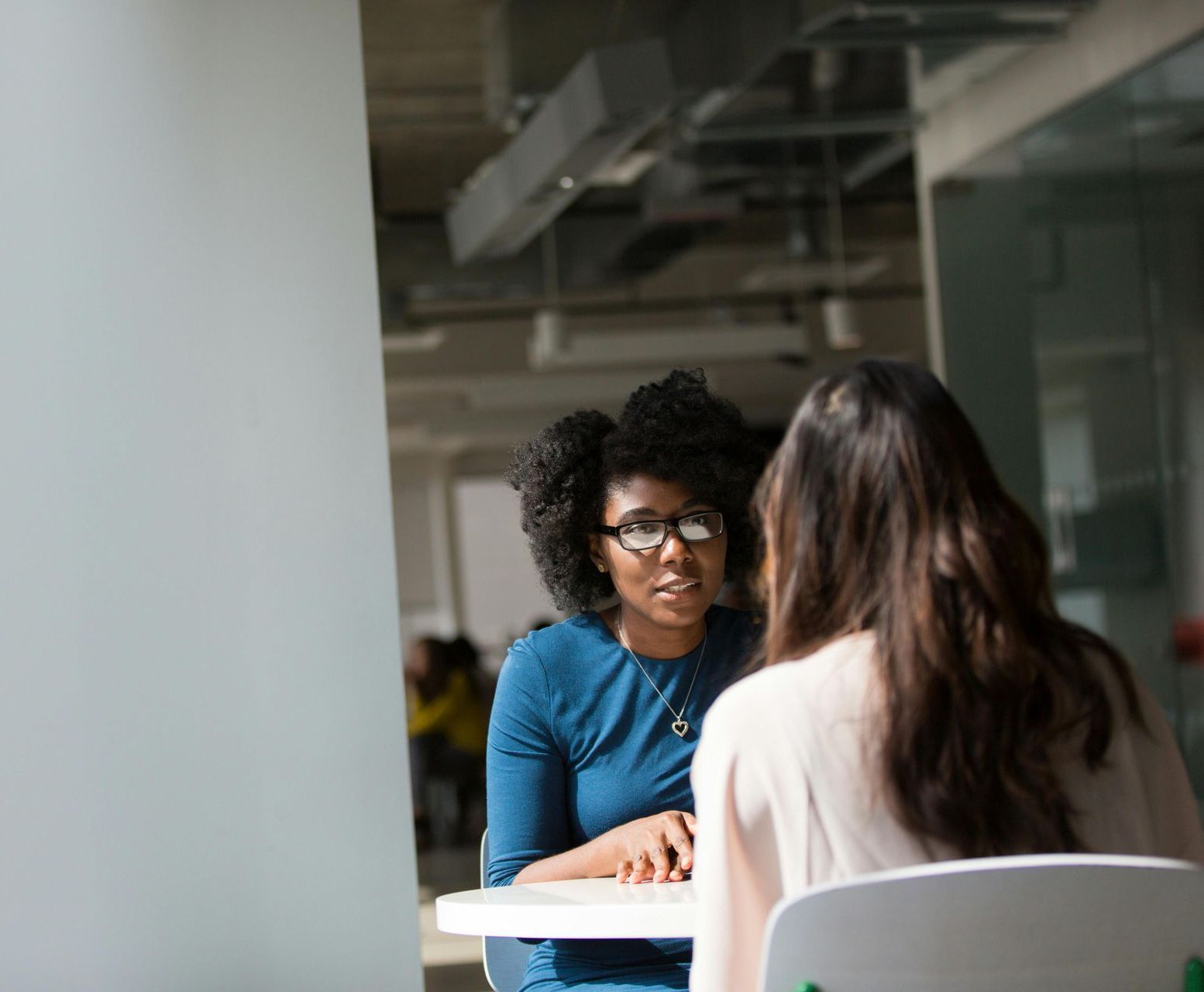Two women seated at a table in an office, one with dark hair and glasses, the other with long hair, engaged in conversation.