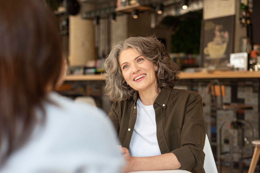 Woman with gray hair smiles at another person in a cafe setting. She is wearing a green jacket over a white shirt.