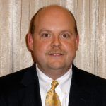 Man in a suit with a mustache, smiling at the camera. He's wearing a gold tie in front of a beige background.