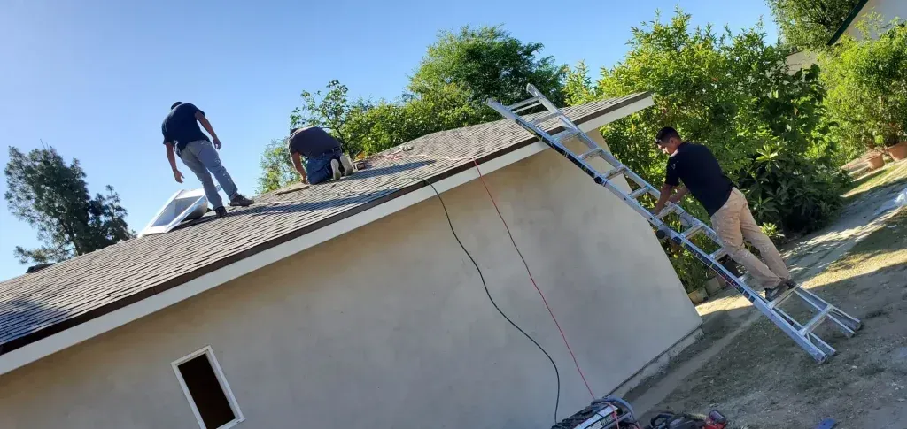Three people on a roof work to secure shingles on a small house.
