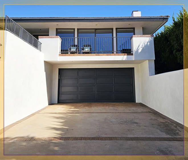 Modern two-story house with a black garage door and balcony, viewed up a concrete driveway