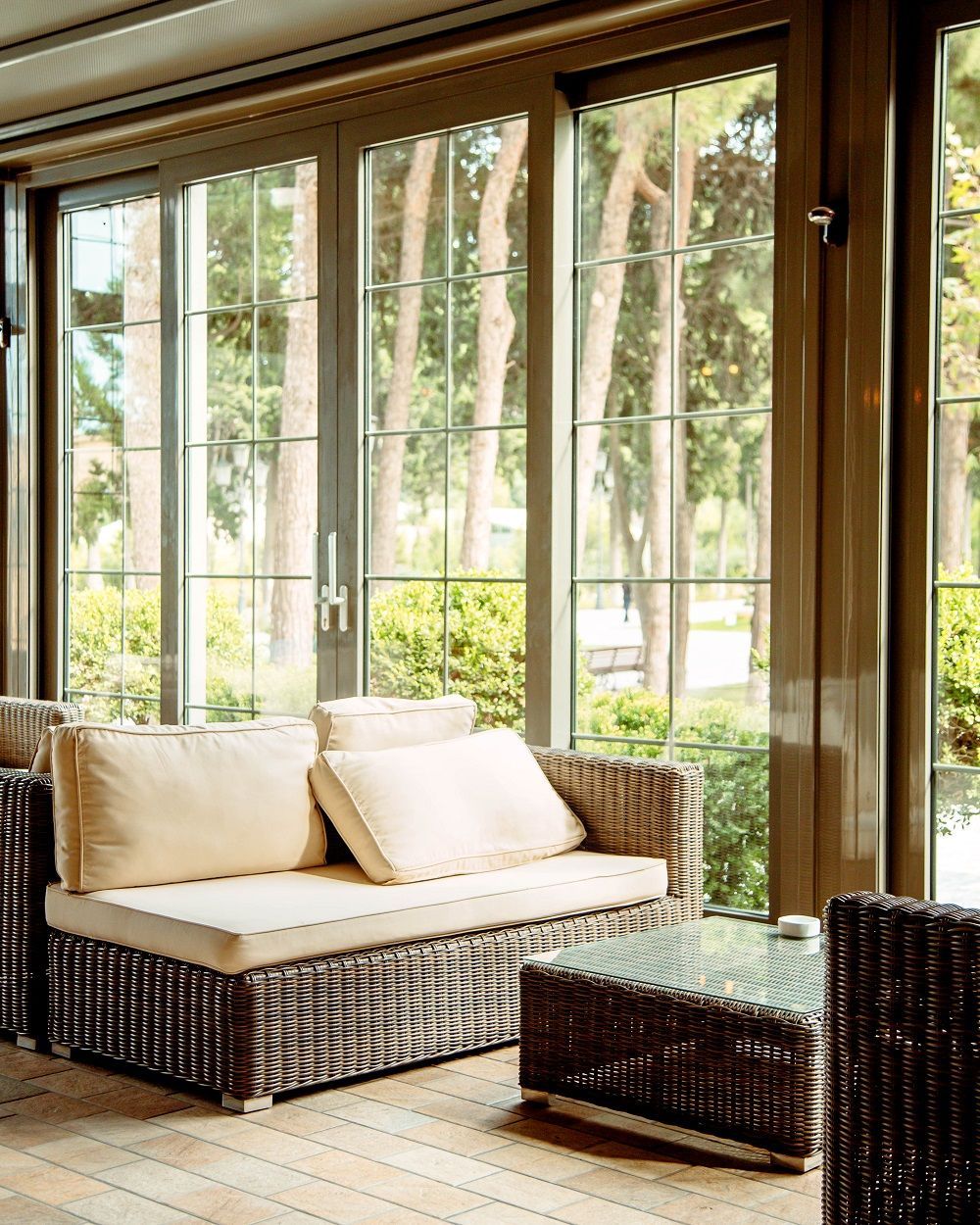 Bright sunroom with wicker sofa, cream cushions, and large windows overlooking greenery
