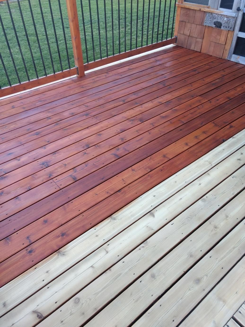 Wooden deck, half stained dark red, other half unfinished. Railing and green lawn in background.