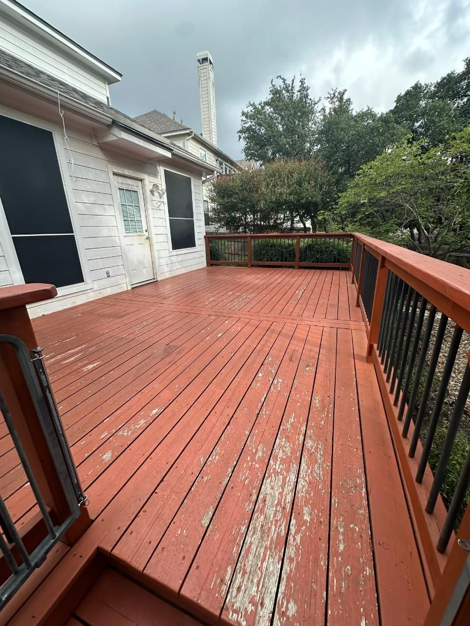 A weathered wooden deck with red stain is attached to a white house. Black railing with trees in the background.